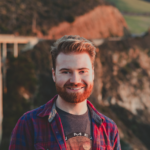 A headshot photograph of Mitchell Lawrence. He has short blond hair and a beard, and is standing in front of an oceanside landscape background.